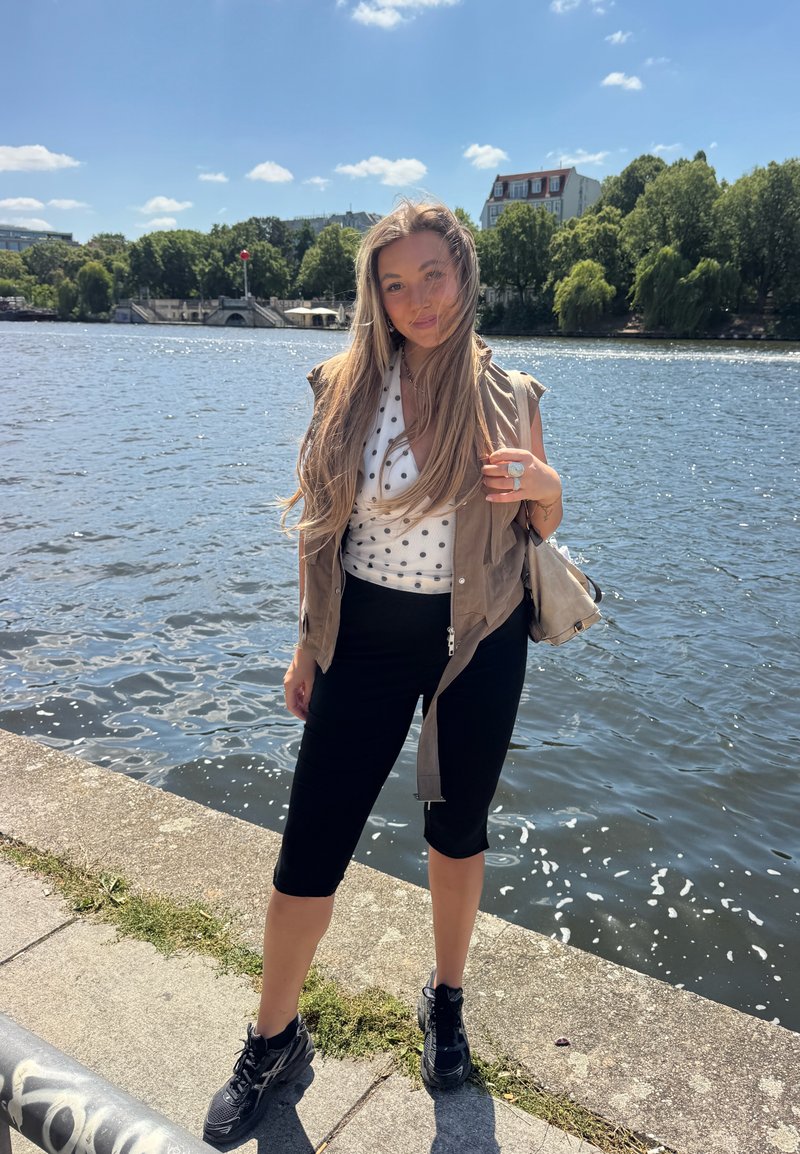 Jeune femme aux longs cheveux se tient près d'une rivière, portant un gilet sans manches, un haut à pois, un pantalon noir raccourci et des baskets noires par une journée ensoleillée.