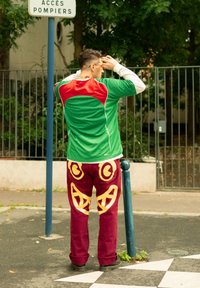 Maillot de sport vert et rouge à manches courtes associé à un pantalon bordeaux avec des motifs abstraits jaunes. Se tenant à côté d'un panneau indicateur.