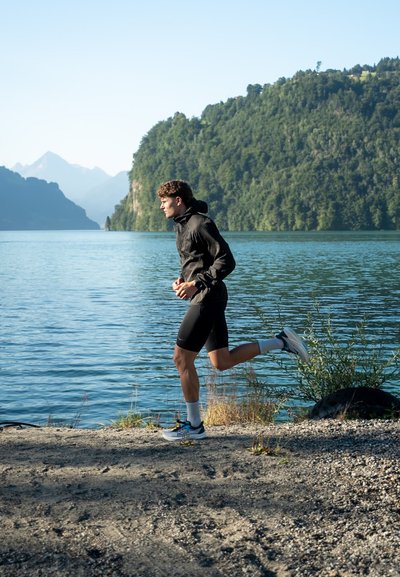 Chaqueta negra impermeable con capucha, pantalones cortos negros ajustados, calcetines blancos y zapatillas de running con acentos de colores, mientras una persona trota junto a un lago.
