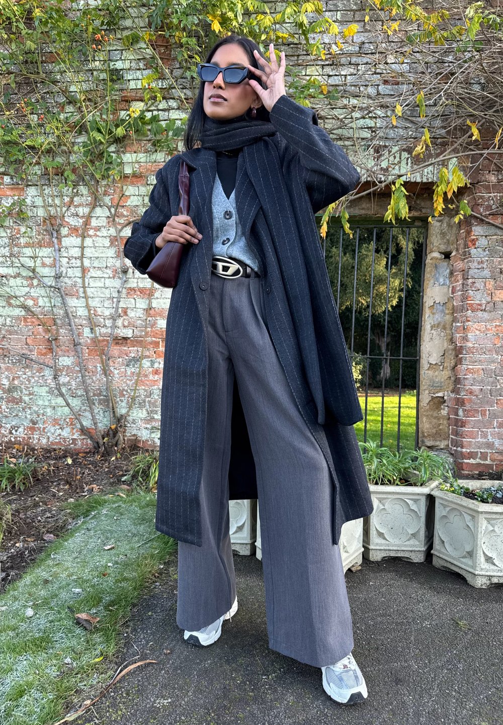 Femme portant de grandes lunettes de soleil noires, une écharpe noire, un long manteau à rayures fines, un pantalon gris, des baskets blanches, et tenant un sac à main marron en extérieur.