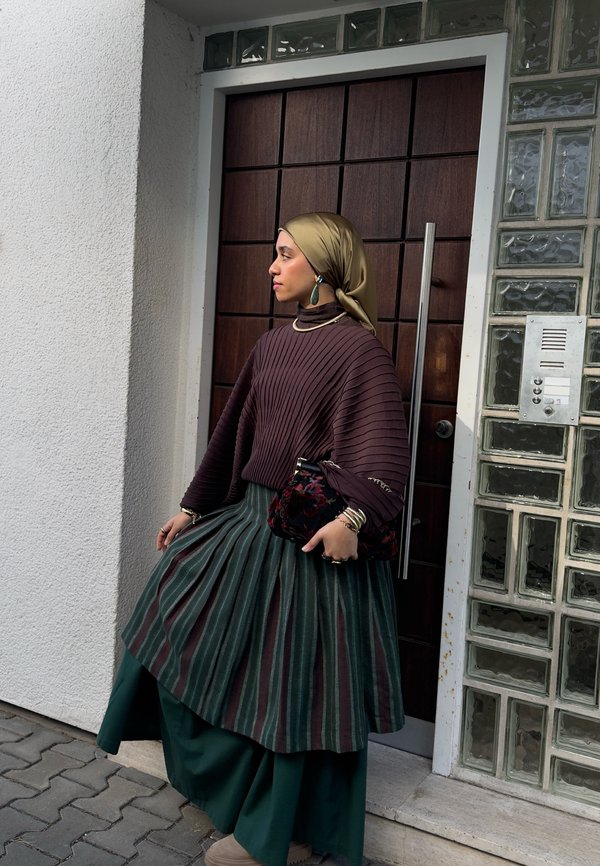 Femme portant un foulard vert, une jupe rayée verte et marron, et un pull texturé marron, debout près d'une porte en bois et d'un mur en blocs de verre.