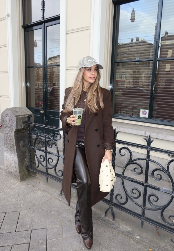 Mujer con abrigo largo marrón, pantalones de cuero y gorra plateada junto a una verja negra de hierro forjado, sosteniendo una bebida verde y un bolso blanco con tachuelas.