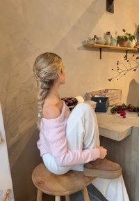Una donna seduta su uno sgabello di legno, indossa un maglione rosa morbido con spalle scoperte e pantaloni bianchi a coste, stivali in stile UGG e una pettinatura con trecce.