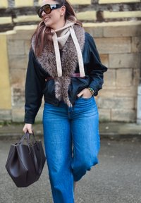 Femme marchant en plein air portant des lunettes de soleil, une veste en cuir noire avec col en fourrure, un jean bleu, et portant un grand sac à main brun foncé.