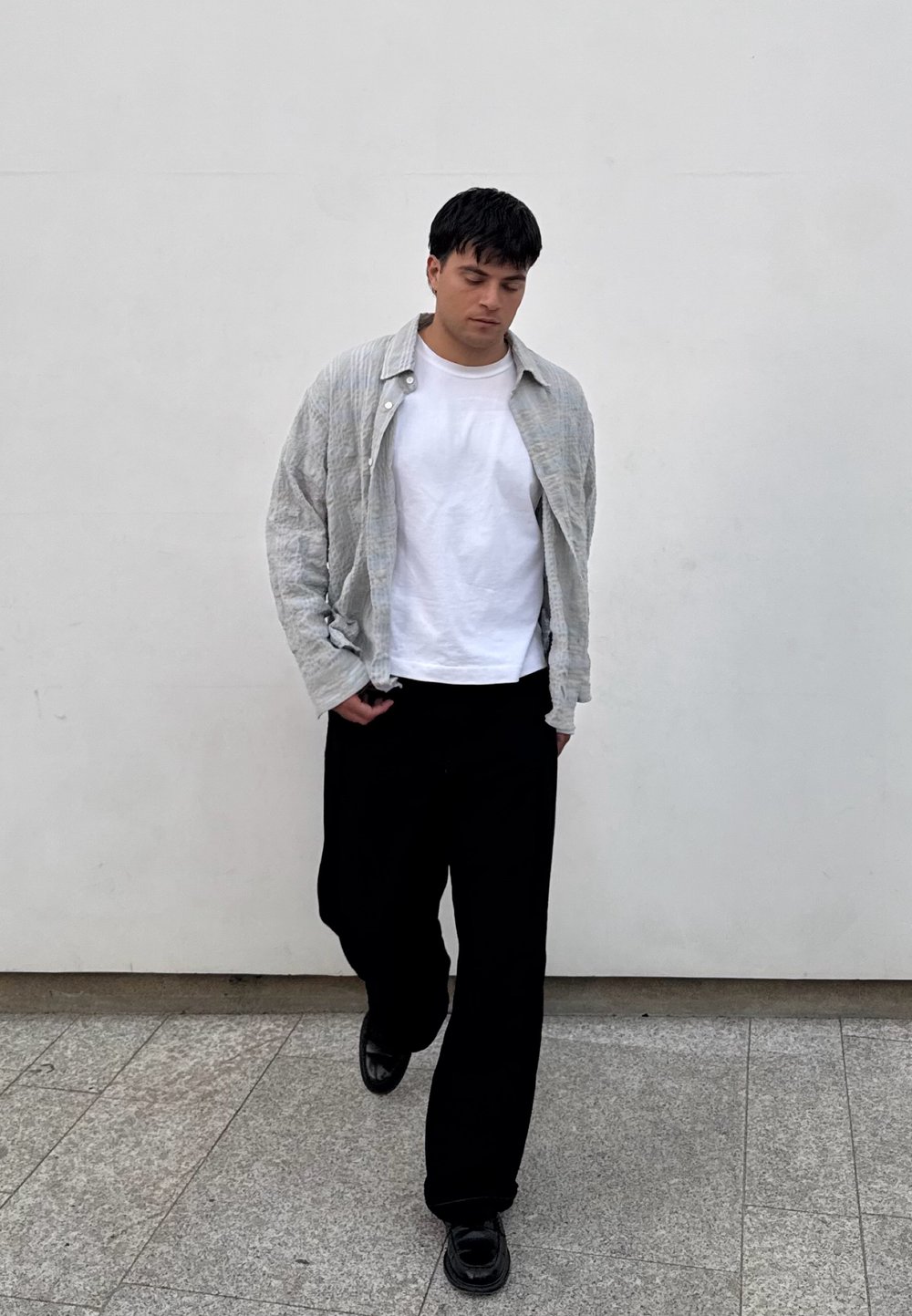 Cămașă oversized texturată de culoare gri deschis peste un tricou alb, asortată cu pantaloni negri largi și pantofi negri lustruiți pe o suprafață de piatră.