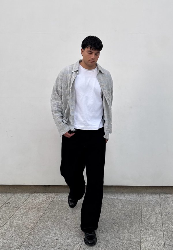 Camicia oversized grigio chiaro con texture sopra una maglietta bianca, abbinata a pantaloni neri larghi e scarpe nere lucide su una superficie di pietra.