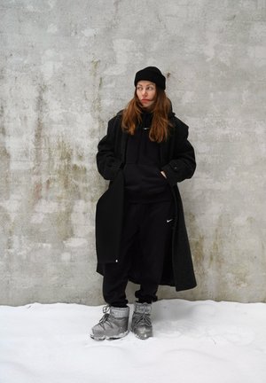 Abrigo de lana negro sobre una sudadera con capucha negra y pantalones de chándal, con botas aislantes grises. El fondo presenta un suelo nevado y una pared gris texturizada.