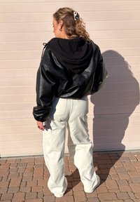 Svart läderjacka i cropped-modell med huva, kombinerad med lösa vita denimbyxor. Skugga kastad på kullerstensytan i solljus.