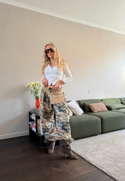 Mujer con cabello largo y rubio que lleva pantalones de camuflaje de gran tamaño, top blanco corto, bolsa beige y botas de tacón alto, de pie en la sala de estar.