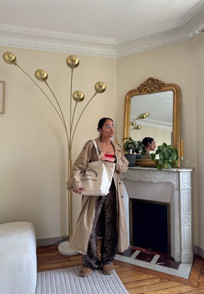 Mujer con abrigo beige tipo trench y pantalones de leopardo junto a una chimenea de mármol con espejo con marco dorado, plantas y una lámpara de pie alta de latón.