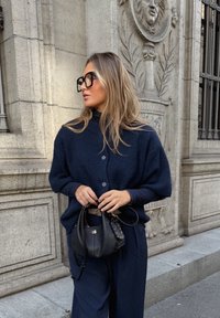 Tengerészkék kötött kardigán túlméretezett fazonnal és gombos záródással, laza tengerészkék nadrággal párosítva. Kiegészítők között egy fekete, texturált kézitáska található.