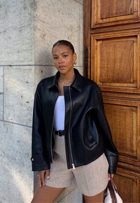 Donna con i capelli slicked-back che indossa una giacca di pelle nera, una maglietta bianca, pantaloni corti beige e orecchini dorati, in piedi accanto a un muro di pietra e a una porta di legno.