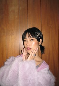 Jeune personne aux cheveux courts noirs, avec des barrettes et des ongles roses, portant un manteau violet duveteux, posant avec les mains près du visage devant un mur en panneaux de bois.