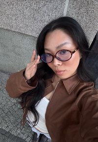 Chaqueta de cuero marrón, camiseta blanca de canalé, gafas de carey redondas con lentes de color púrpura claro, cabello largo y negro, de pie frente a un fondo texturizado.