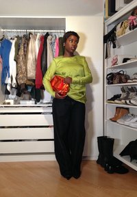 Blusa verde de cetim de mangas longas, jeans escuros, segurando uma carteira metálica laranja. O fundo apresenta diversas roupas e sapatos em prateleiras.