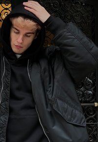 Jeune personne aux cheveux décolorés portant un sweat à capuche noir et une veste, ajustant sa capuche, debout devant une porte en fer forgé décorative.