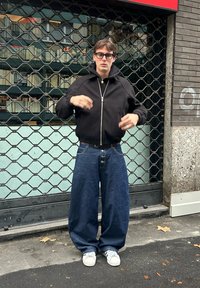 Ung mann iført store blå jeans, svart jakke med glidelås, hvite joggesko og briller står på en bygate foran en metallport.