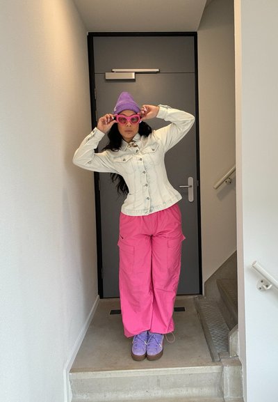 Chaqueta de mezclilla blanca con cuello puntiagudo, pantalones cargo rosas, gorro lavanda, gafas de sol rosas de gran tamaño y zapatillas moradas, frente a una puerta gris.