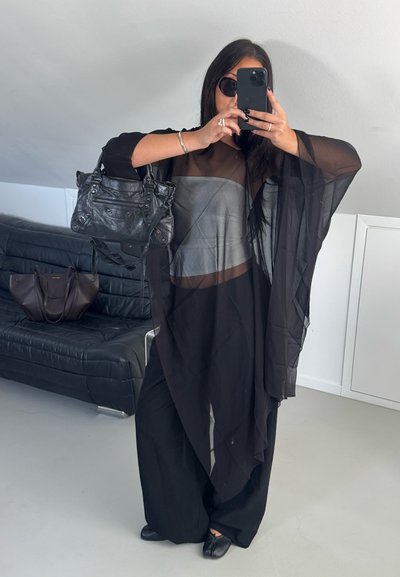 Mujer con top negro transparente, pantalones anchos negros y gafas de sol, sosteniendo un teléfono para selfie en el espejo y llevando un bolso negro.