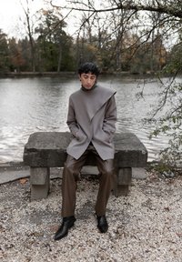 Person i grå asymmetrisk frakke og brune bukser sidder på en stenbænk ved en sø under bare trægrene om efteråret.