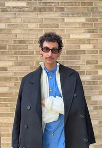 Uomo con capelli ricci e baffi indossa occhiali da sole neri, un cappotto nero sopra una giacca bianca e una camicia blu con decorazioni argentate, davanti a un muro di mattoni.