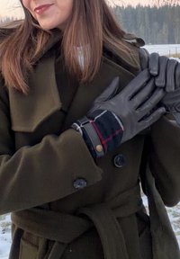 Γυναίκα έξω φορώντας σκούρο πράσινο παλτό και μαύρα δερμάτινα γάντια με καρό μανσέτες, προσαρμόζοντας ένα γάντι μπροστά σε ένα χιονισμένο, δασώδες τοπίο.