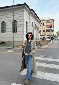 Rutigt trench coat, grå väst, vit skjorta och blå jeans. Bruna ankelskor och solglasögon. Stadsmiljö med byggnader och övergångsställe.