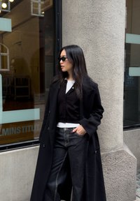 Manteau en laine noire porté sur un cardigan noir, une chemise blanche et un jean large en denim foncé. Accessoires comprenant des lunettes de soleil noires. Fond texturé.