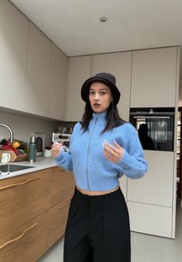 Pull court bleu clair avec une fermeture éclair, en tissu doux ; associé à un pantalon noir ample. Chapeau bob marron et bijoux simples.