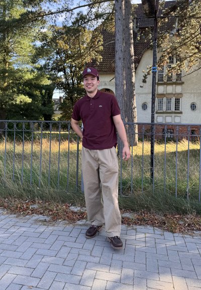 Camisa polo granate, pantalones beis y zapatos marrón oscuro. De pie en un camino de piedras cerca de una valla y vegetación bajo un cielo despejado.