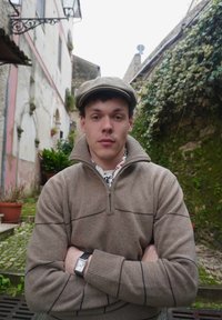 Jeune homme en pull beige et casquette, les bras croisés, debout dans une ruelle étroite pavée avec des plantes et des murs couverts de lierre.