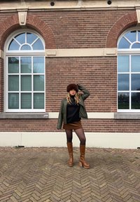 Bruine bonten muts, groene leren jas, zwarte coltrui, bruine suède rok, zwarte panty's en tan hoge laarzen met hakken tegen een bakstenen muur.