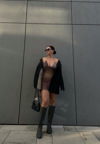 Vestido marrón con escote halter y patrón degradado, blazer negro oversized, botas oscuras hasta la rodilla y un pequeño bolso negro contra una pared gris.