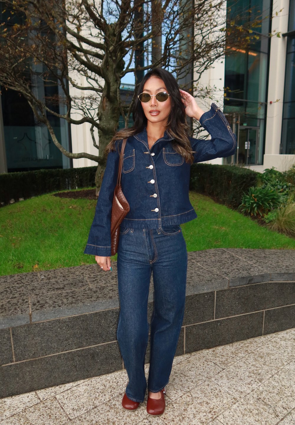 Giubbotto di jeans con bottoni e due tasche sul petto, abbinato a pantaloni in denim aderenti. Scarpe in pelle marrone e una borsa a tracolla marrone. Occhiali da sole circolari.