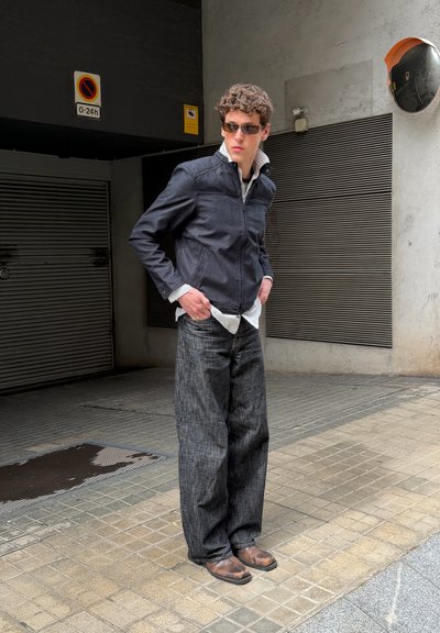 Joven con gafas de sol, chaqueta negra, camisa de rayas, pantalones anchos y oscuros y zapatos marrones, de pie en una acera de baldosas cerca de puertas de garaje.