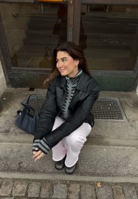 Veste en cuir noire sur un pull à rayures grises, pantalon rose clair, bottes noires, assise sur des marches avec un sac bleu marine à côté d'elle.