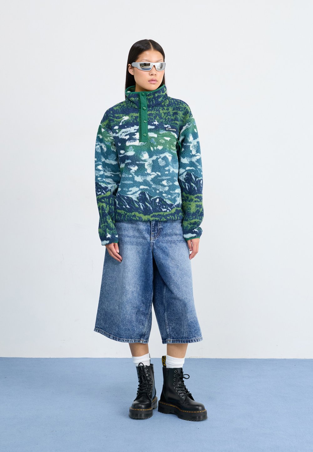 Fleece pullover in a blue and green mountain pattern, featuring a high collar and button placket, paired with wide denim shorts and black boots.