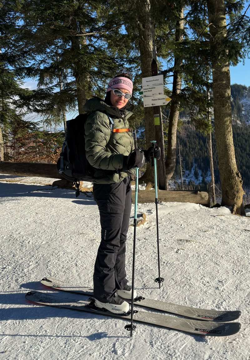 Personne vêtue d'équipements d'hiver et de lunettes de soleil, debout sur des skis avec des bâtons près d'un arbre portant un panneau indicateur multi-directionnel dans une forêt enneigée.