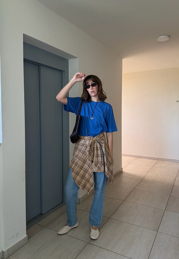 Mujer con gafas de sol, camisa azul, camisa de cuadros atada alrededor de la cintura, vaqueros claros y zapatos beige, parada cerca de las puertas cerradas del ascensor en el interior.