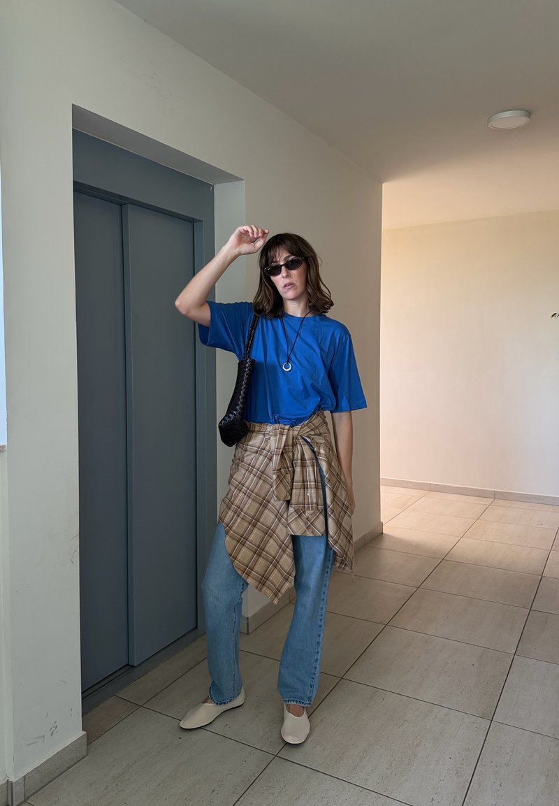 Femme portant des lunettes de soleil, une chemise bleue, une chemise à carreaux nouée autour de la taille, un jean clair et des chaussures beige se tient près des portes fermées de l'ascenseur à l'intérieur.