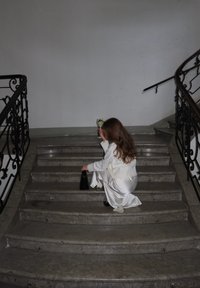 Ένα άτομο ντυμένο στα λευκά κάθεται σε μαρμάρινες σκάλες, κρατώντας μια μαύρη τσάντα και ένα ποτήρι με ποτό, με μια διακοσμητική μεταλλική ράγα κοντά.