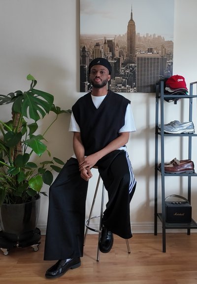 Chaleco negro sobre camiseta blanca, pantalones anchos negros, zapatos negros. Ambiente interior con una planta, un taburete y una estantería que muestra zapatos y un altavoz.