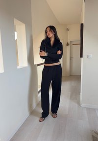 Cardigan negro con cuello de piel sintética, combinado con pantalones negros sueltos y sandalias con tiras. Accesorios simples completan el look.