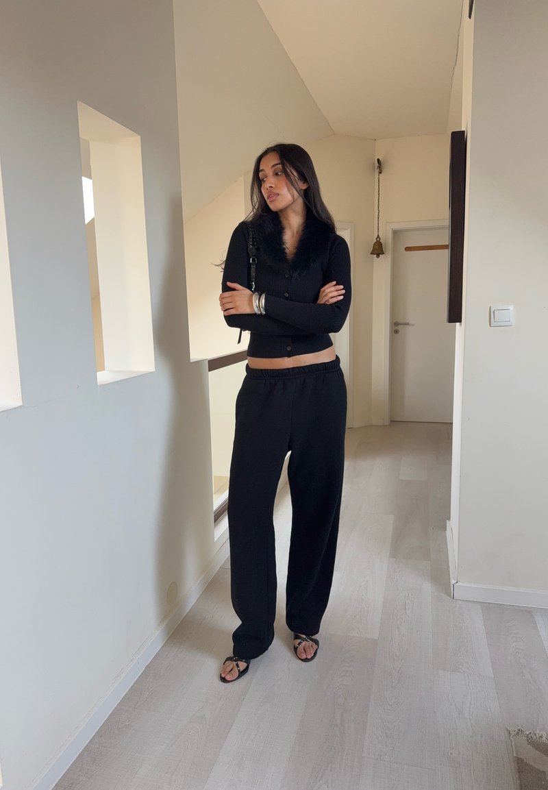 Cardigan noir avec col en fausse fourrure, associé à un pantalon noir ample et des sandales à lanières. Des accessoires simples complètent le look.