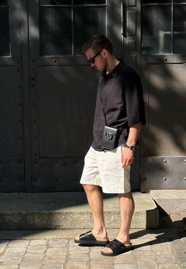 Homme portant une chemise noire, un short blanc, des sandales noires et des lunettes de soleil marchant devant une grande porte métallique sombre à la lumière du soleil et à l'ombre.