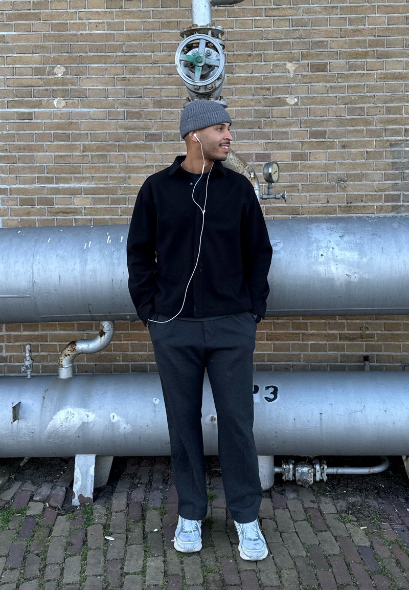 Homme portant un bonnet gris, une veste noire et un pantalon gris, les mains dans les poches, écoutant des écouteurs, debout devant des tuyaux métalliques sur un mur en briques.