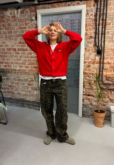 Cárdigan rojo con botones, camiseta blanca debajo y pantalones verde con estampado de leopardo. Zapatillas grises sobre un suelo de hormigón contra una pared de ladrillo.