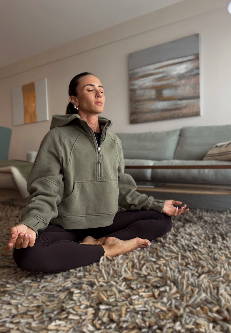Oversized Hoodie in olivgrün mit halbem Reißverschluss, entspannter Passform und großer Fronttasche. Sitzt auf einem strukturierten Teppich mit barfuß in Meditationshaltung.