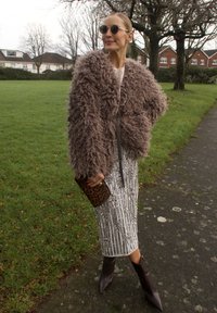 Femme portant un manteau brun duveteux, une jupe argentée à sequins, des bottes brunes pointues, des lunettes de soleil rondes, tenant une pochette à imprimé léopard, debout sur un sentier près de l'herbe.