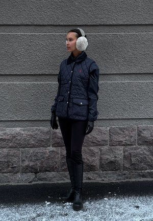 Chaqueta acolchada azul marino con bolsillos con botones, usada con guantes negros y botas altas. El modelo lleva orejeras blancas de peluche y está de pie sobre un pavimento nevado.